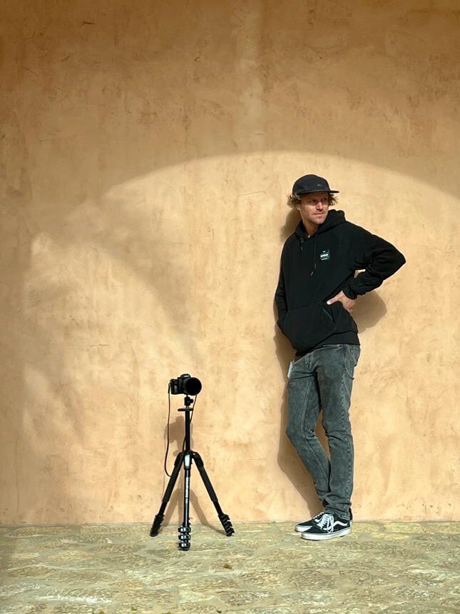 Chris Gollhofer steht vor einer Wand, seine Kamera steht neben ihm auf einem Stativ - Chris Gollhofer Lifestylefotograf - Spanien